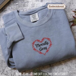 A gray sweatshirt with a red heart-shaped embroidered on it, featuring the text "Morally Grey" in a cursive font. The sweatshirt is folded and placed on a gray surface, with a leaf and a book visible in the background.