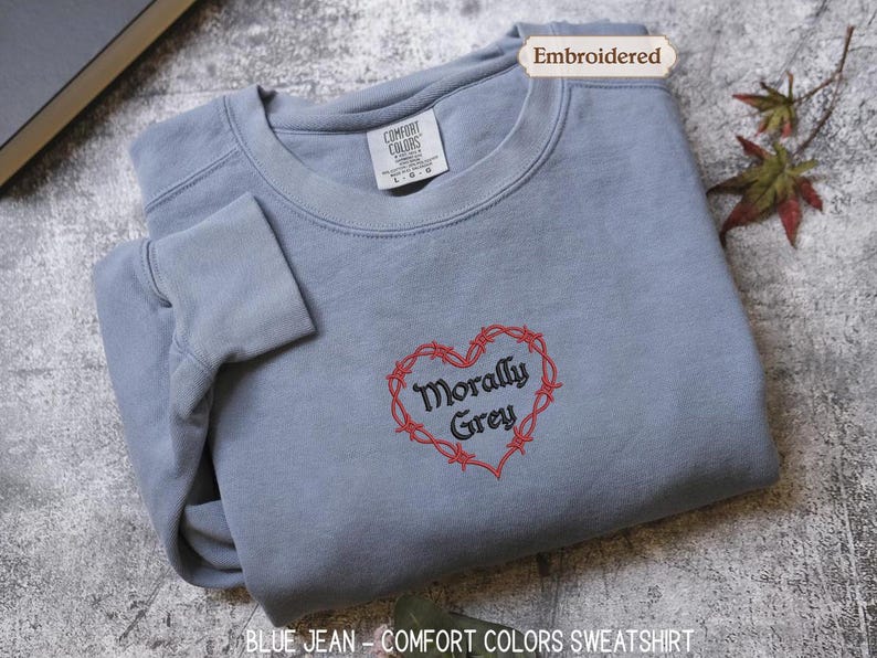 A gray sweatshirt with a red heart-shaped embroidered on it, featuring the text "Morally Grey" in a cursive font. The sweatshirt is folded and placed on a gray surface, with a leaf and a book visible in the background.
