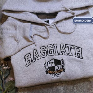 A grey hoodie with the word "BASGIAH" and a crest embroidered on it, along with some leaves and flowers.