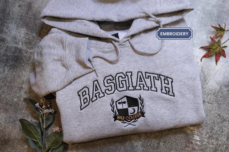 A grey hoodie with the word "BASGIAH" and a crest embroidered on it, along with some leaves and flowers.