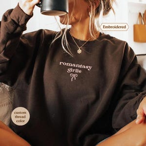 A woman wearing a brown sweater with the text "romantic girl" printed on it, sitting and drinking from a mug.
