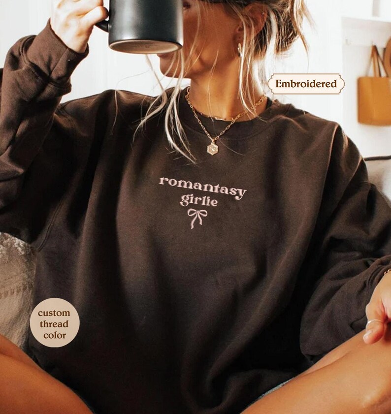 A woman wearing a brown sweater with the text "romantic girl" printed on it, sitting and drinking from a mug.