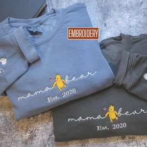 May include: Two sweatshirts, one blue and one grey, with embroidered designs. The blue sweatshirt has "mama bear" with a Winnie the Pooh graphic and "Est. 2020". The grey sweatshirt has the same design. Sleeves have names.