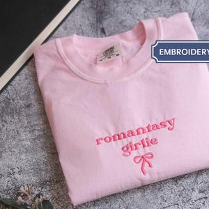 A pink t-shirt with the text "romantic girl" printed on it, along with a bow, is folded and placed on a gray surface next to a black book and some leaves.