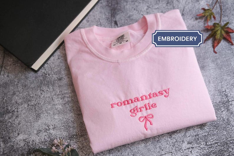 A pink t-shirt with the text "romantic girl" printed on it, along with a bow, is folded and placed on a gray surface next to a black book and some leaves.