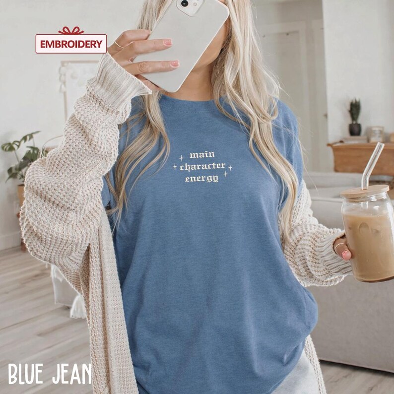 A woman wearing a blue t-shirt with the text "main character energy" printed on it, holding a smartphone and a glass of iced coffee.