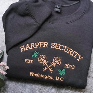 A black sweatshirt with embroidered text and graphics, including a key design, is displayed on a gray surface alongside a small plant with white flowers.