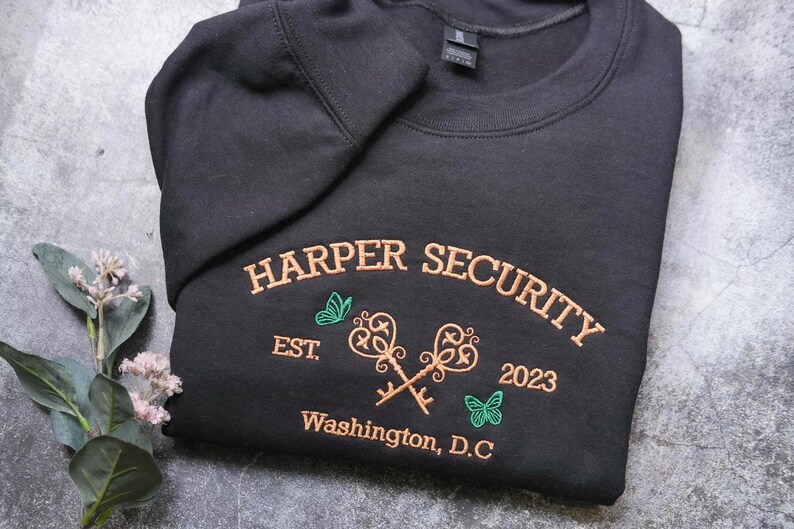 A black sweatshirt with embroidered text and graphics, including a key design, is displayed on a gray surface alongside a small plant with white flowers.