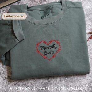a gray sweatshirt with a red heart-shaped embroidered design and the text "Morally Grey" printed on it. The sweatshirt is folded and placed on a gray surface, with a leaf visible in the background.