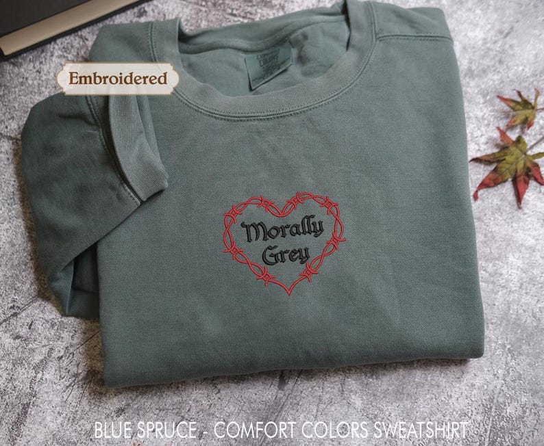 a gray sweatshirt with a red heart-shaped embroidered design and the text "Morally Grey" printed on it. The sweatshirt is folded and placed on a gray surface, with a leaf visible in the background.