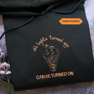 May include: Dark green hoodie with embroidered text and a lightbulb design. The text reads "all lights turned off CAN BE TURNED ON". The word "EMBROIDERED" is on an orange tag. A sprig of pink flowers is in the corner.