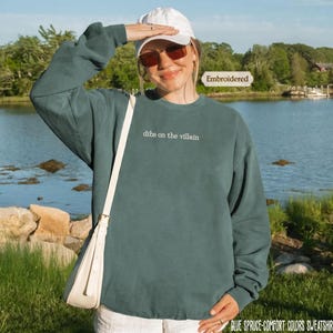 A woman wearing a green sweatshirt with the text "dibs on the villain" stands in front of a body of water, smiling and holding her hand up to her face.