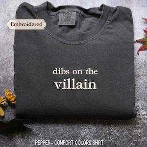 May include: A charcoal grey crewneck t-shirt featuring the white embroidered text "dibs on the villain" centred on the chest. The garment includes a Comfort Colors brand label at the inner neckline indicating a size 2XL.