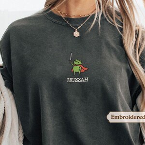 A person wearing a gray sweatshirt with a green frog graphic and the word "HUZZAH" printed on it.