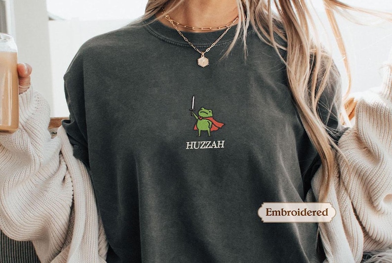 A person wearing a gray sweatshirt with a green frog graphic and the word "HUZZAH" printed on it.