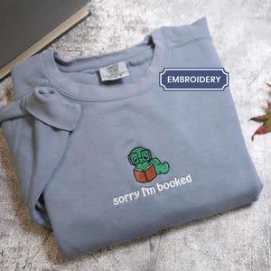 Peut inclure: Un sweat-shirt bleu clair avec une broderie d'un ver de livre vert portant des lunettes et lisant un livre. Le texte "sorry I'm booked" est brodé en dessous. Le sweat-shirt est plié sur une surface grise.