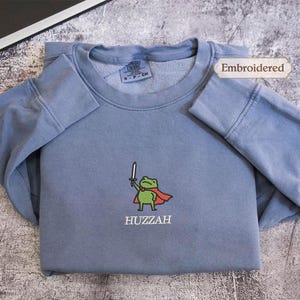 A gray sweatshirt with a frog character and the word "HUZZAH" printed on it, along with a label indicating that it is embroidered.