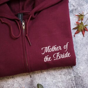 A burgundy zip-up hoodie with the text "Mother of the Bride" printed on it, along with a small leaf and a plant.