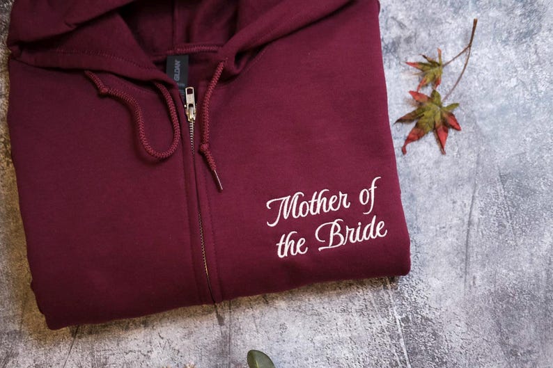 A burgundy zip-up hoodie with the text "Mother of the Bride" printed on it, along with a small leaf and a plant.