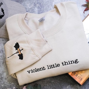 a beige sweatshirt with a black dragon embroidered on the sleeve, along with a gray baseball cap and a book titled "The Earth" by Rosario.
