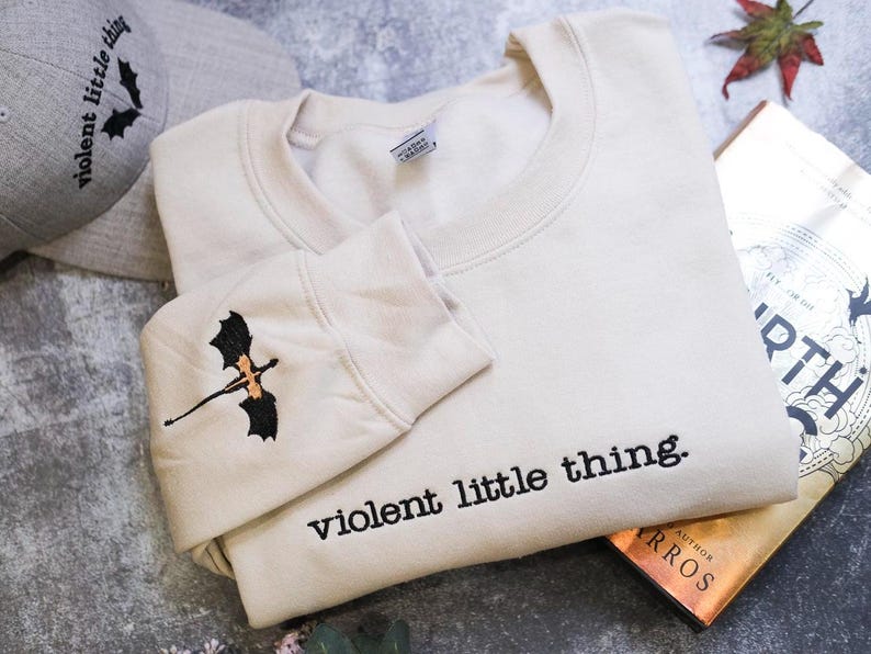 a beige sweatshirt with a black dragon embroidered on the sleeve, along with a gray baseball cap and a book titled "The Earth" by Rosario.