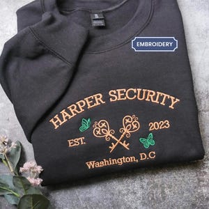 A black sweatshirt with a blue embroidered label that says "EMBROYERY" and features text and images related to "HARPER SECURITY" and "Est. 2023 Washington, D.C."