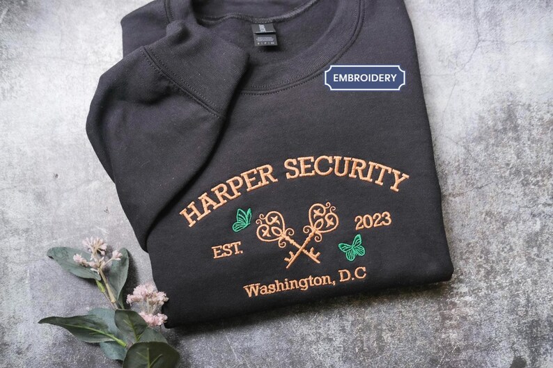 A black sweatshirt with a blue embroidered label that says "EMBROYERY" and features text and images related to "HARPER SECURITY" and "Est. 2023 Washington, D.C."