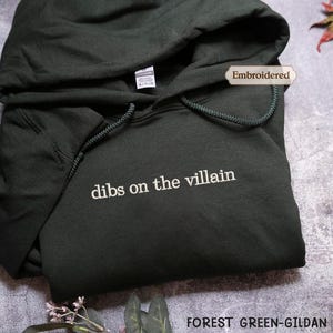 A dark green hoodie with the text "dibs on the villain" printed on it, along with a label that says "Embroidered". The hoodie is placed on a gray surface, and there is a leaf and some books in the background.