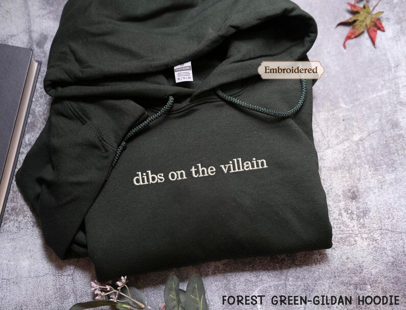 A dark green hoodie with the text "dibs on the villain" printed on it, along with a label that says "Embroidered". The hoodie is placed on a gray surface, and there is a leaf and some books in the background.
