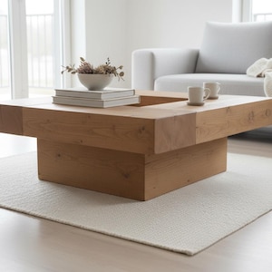 Peut inclure: Une table basse en bois brun clair de forme carrée avec un centre en retrait. La table a une base empilée et est placée sur un tapis blanc. Deux petites tasses blanches et un bol avec des fleurs séchées sont sur la table.