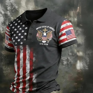 Puede incluir: Un polo gris con un diseño desgastado de la bandera estadounidense. El lado izquierdo muestra las estrellas y las rayas, mientras que la manga derecha tiene una pequeña bandera. La camisa incluye un emblema de águila y el texto "UNITED STATES OF AMERICA 1776-2026".