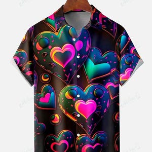 May include: Short-sleeved button-down shirt with a vibrant heart pattern. The design features neon-colored hearts in shades of pink, blue, and green against a black background. The shirt has a classic collar and white short sleeves.