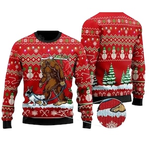 May include: A red long-sleeve crewneck sweater featuring a graphic of Bigfoot wearing a Santa hat and carrying a gift sack while walking a French Bulldog. The sweater has a repeating pattern of snowmen, Christmas trees, and string lights.