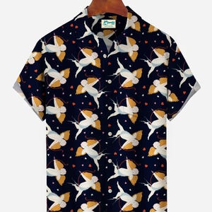 Op de afbeelding: Marineblauwe shirt met korte mouwen met een herhalend patroon van witte cherubijnen met gele vleugels, bogen en pijlen en kleine rode hartjes. De shirt heeft een klassieke kraag en hangt aan een houten kleerhanger.