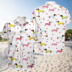 May include: White button-down shirt featuring a repeating pattern of dachshunds wearing various outfits, including sweaters and coats. The dogs are in yellow, pink, blue, and green, with pink hearts scattered throughout the design. The shirt has short sleeves.
