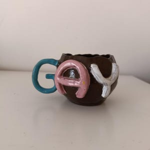 Puede incluir: Una taza de cerámica hecha a mano con la palabra "GAY" en letras en relieve. La taza es marrón con un asa azul. Las letras son rosas y blancas.