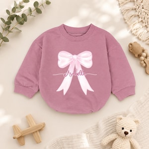 May include: A mauve long-sleeve sweatshirt featuring a large, light pink illustrated bow graphic centered on the chest. The name Arabella is printed in a cursive script across the center of the bow. The garment has a ribbed crew neckline.