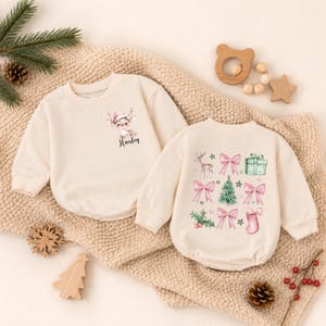 May include: Two cream-colored long-sleeve sweatshirts. The left one features a small deer wearing a pink hat and the name Haisley. The right one displays a pattern of deer, pink bows, a green gift, a Christmas tree, holly, and a pink stocking.