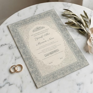 May include: A decorative wedding certificate with a grey and cream design. The names Yusuf Alvi and Mariam Noor are featured on the certificate. Two gold wedding rings and a sprig of greenery with a ribbon are also visible.