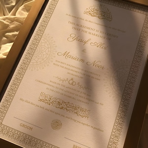 May include: A framed marriage certificate with gold accents. The document features ornate calligraphy and the names Yusuf Alvi and Mariam Noor. The frame is a warm gold color, and the text is in English and Arabic.