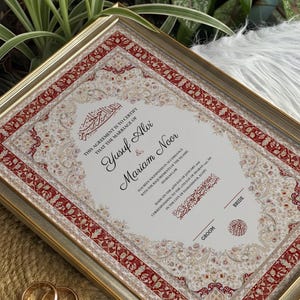 May include: A framed Islamic marriage certificate with a red and cream ornate border. The text reads: This agreement is to certify that the marriage of Yusuf Alvi & Mariam Noor has been solemnized in accordance with the requirements of the Islamic Shariah Law.