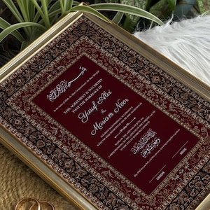 Pode incluir: Um certificado de casamento emoldurado com borda dourada, apresentando um design estampado em bordô e preto. O certificado inclui os nomes Yusuf Alvi e Mariam Noor, com escrita árabe e texto em inglês. Dois anéis de casamento dourados são visíveis.