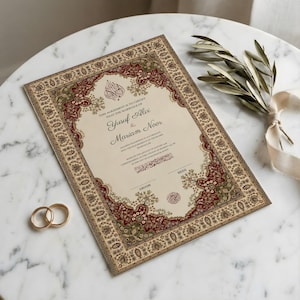 May include: A decorative wedding certificate with a beige background and intricate red and green borders. The names "Yusuf Alvi & Mariam Noor" are displayed. Two gold wedding rings are placed near the document.
