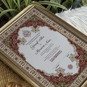 May include: A framed marriage certificate with a gold border. The document includes the names Yusuf Alvi and Mariam Noor, with decorative red, green, and brown borders. Two gold wedding rings are in the foreground.