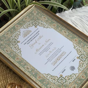 May include: A framed Islamic marriage certificate featuring a decorative border with intricate green, cream, and orange floral patterns. The document text includes the names Yusuf Alvi and Mariam Noor, dated June 2025, with two gold wedding bands nearby.