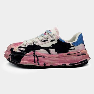 Op de afbeelding: Een unieke, artistieke sneaker met een dikke, getextureerde zool. De schoen heeft een wit bovenwerk met roze en zwarte verfspatten. De hiel heeft een blauw accent en de veters zijn wit met kleurrijke uiteinden. De zool is ongeveer 5 cm dik.