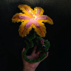 Può includere: Un'artigianato fatto a mano e stravagante con un grande fiore giallo e rosa con uno stelo verde e un camaleonte verde. Il fiore ha piccole luci al centro. Il camaleonte ha grandi occhi neri e un fiore rosa sulla testa.