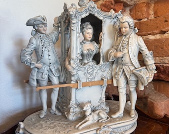 May include: A detailed porcelain figurine depicting a carriage scene with three figures in 18th-century attire. The carriage is ornate, and a small dog rests at the base. The color palette is primarily off-white with subtle gray accents.