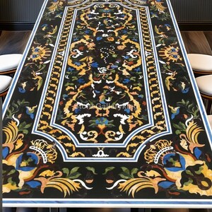 Könnte beinhalten: Ein rechteckiger Tisch aus schwarzem Marmor mit einem aufwendigen eingelegten Blumendesign in Gelb-, Blau-, Weiß- und Orangetönen. Der Tisch hat einen weißen Rand mit blauem Akzent. Er steht auf einem dunklen Holzboden.