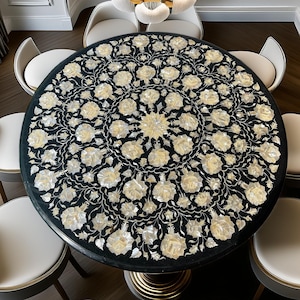 Könnte beinhalten: Ein runder, schwarzer Esstisch mit einem floralen Intarsienmuster in Weiß und Creme. Der Tisch wird von einem goldfarbenen Sockel getragen. Acht weiße Stühle mit geschwungenen Rücken umgeben den Tisch.